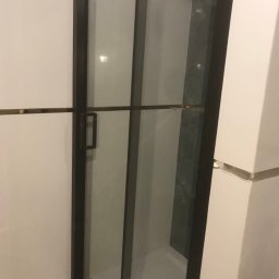 Norbert - Nowoczesna kabina prysznicowa z czarnymi profilami i szklanymi drzwiami, widoczny fragment szarej ściany wewnątrz kabiny, białe płytki na ścianach zewnętrznych.