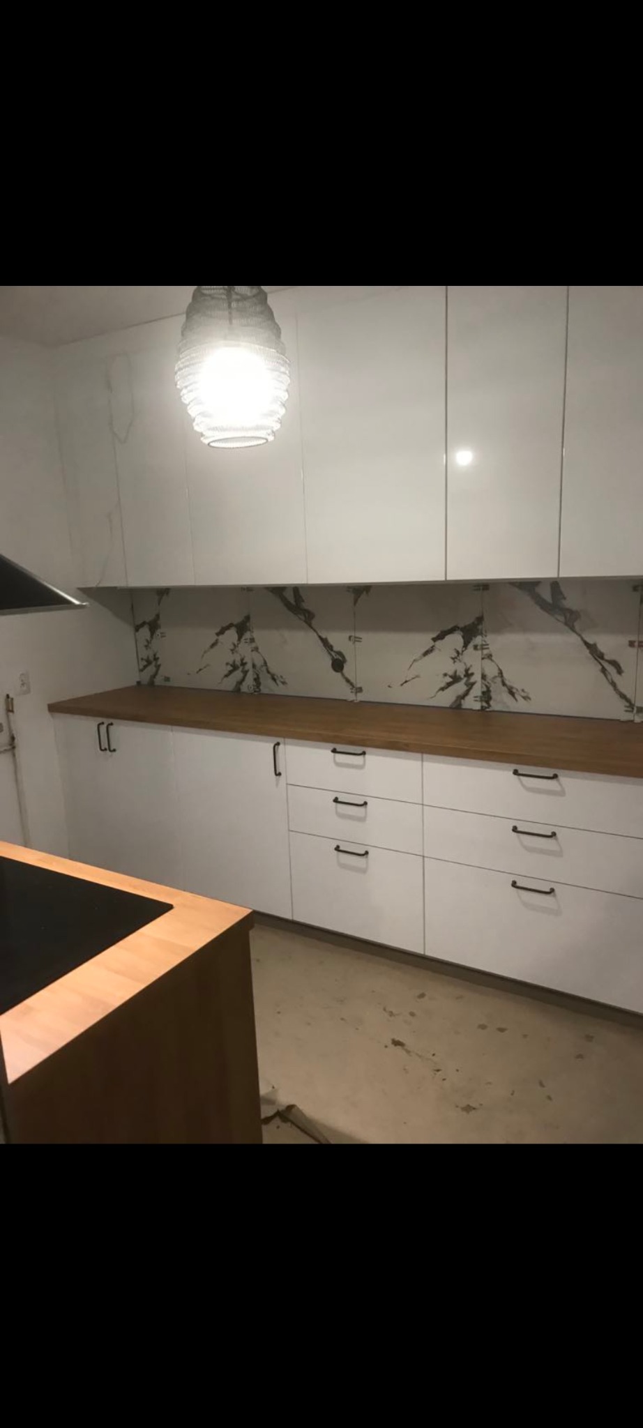 Białe szafki kuchenne z drewnianym blatem i marmurowym panelem ściennym, oświetlone wiszącą lampą z plecionki, na podłodze widoczne ślady po remoncie.