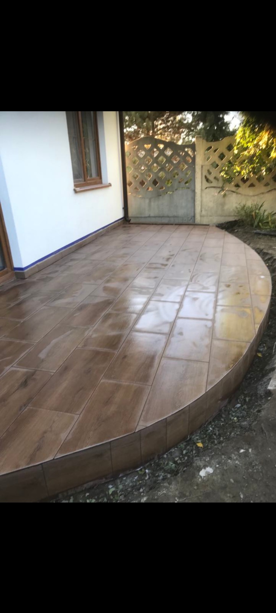 Wyłożony brązowymi płytkami taras przylegający do białego budynku z oknem, widoczne ogrodzenie z ażurowym wzorem.