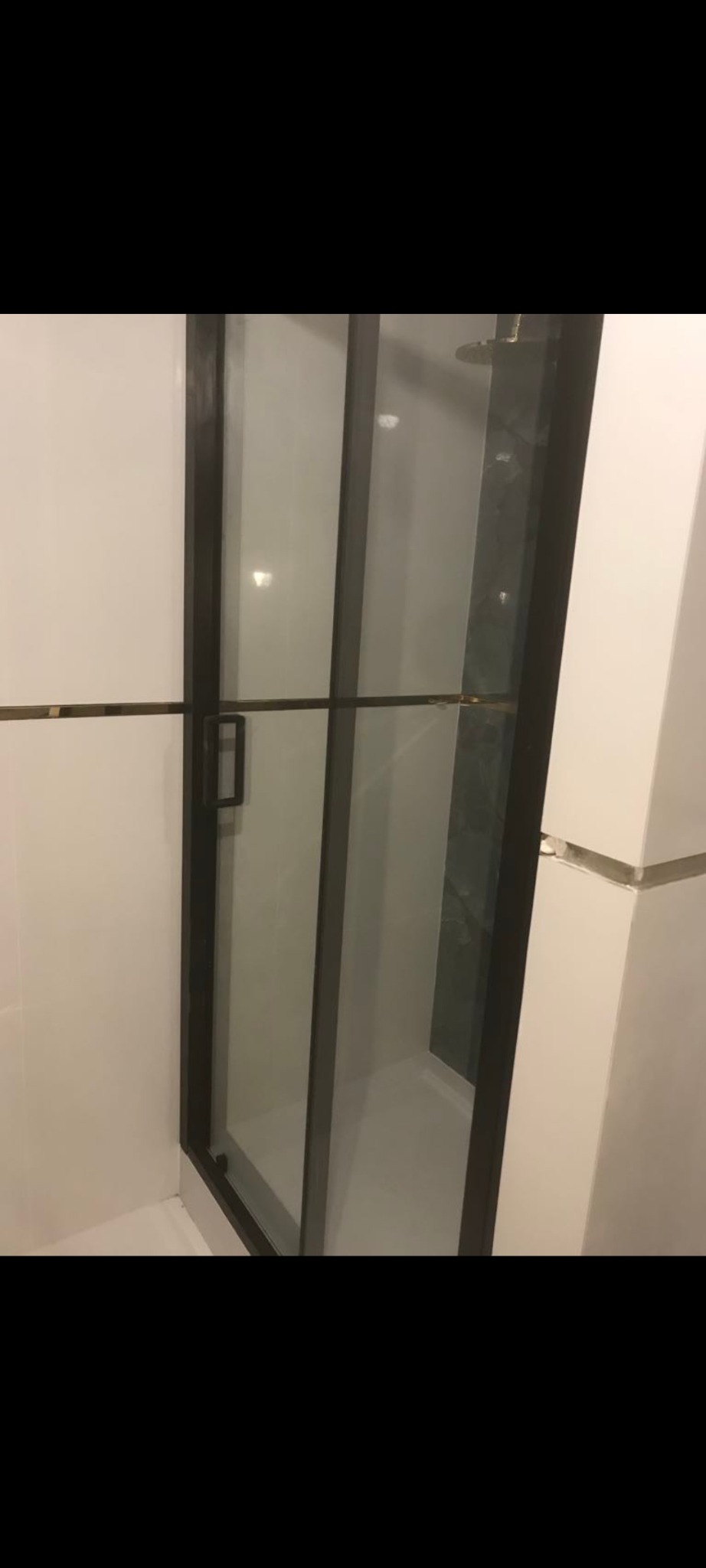 Nowoczesna kabina prysznicowa z czarnymi profilami i szklanymi drzwiami, widoczny fragment szarej ściany wewnątrz kabiny, białe płytki na ścianach zewnętrznych.