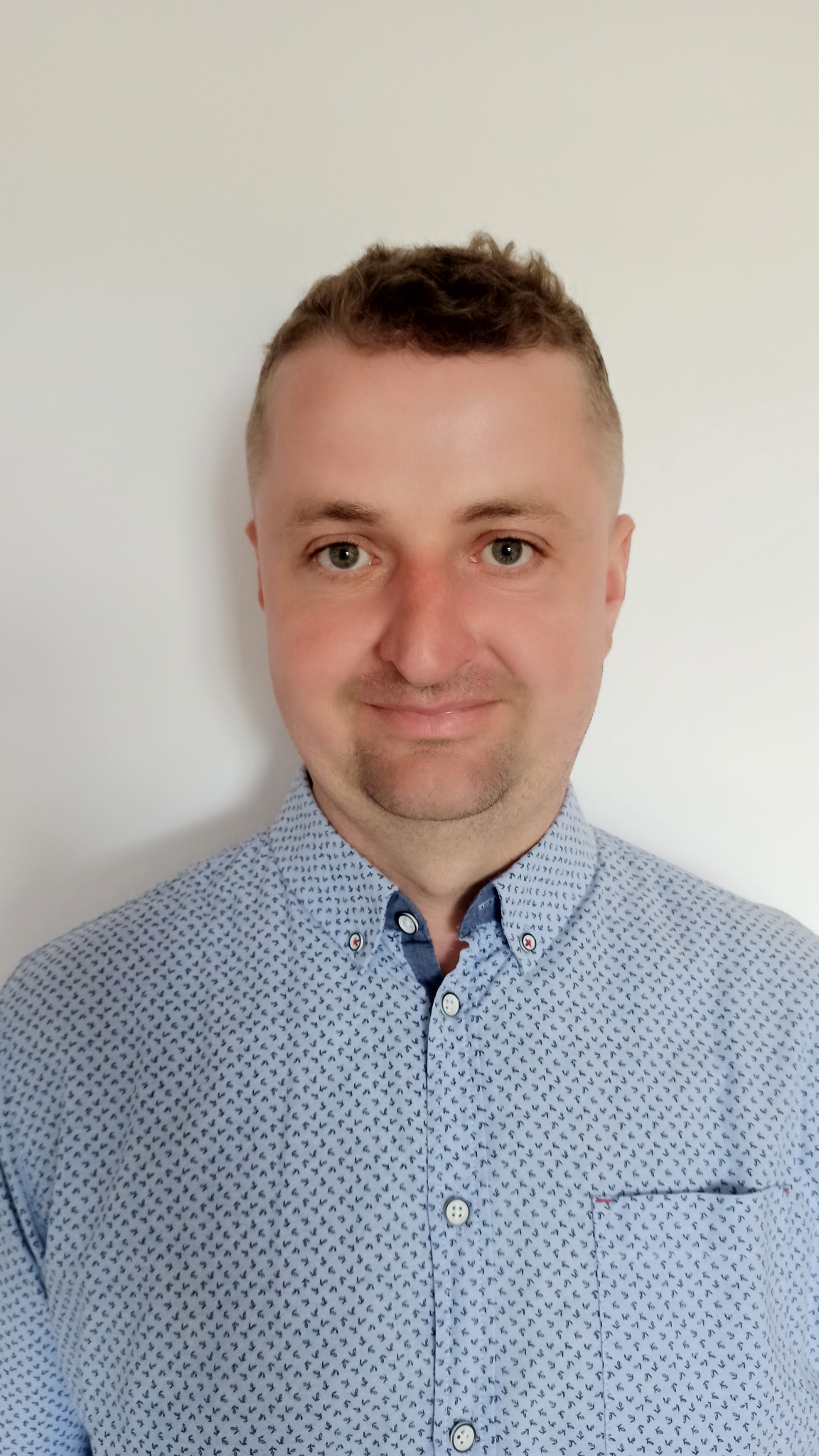 Portret uśmiechniętego mężczyzny w błękitnej koszuli z drobnym wzorem i kieszenią, na jednolitym białym tle.