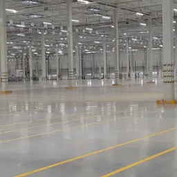 Przestronne, puste wnętrze magazynu z polerowaną posadzką, żółtymi liniami wyznaczającymi strefy, wysokim sufitem z oświetleniem i rzędami kolumn z czarno-żółtymi pasami ostrzegawczymi u podstawy.