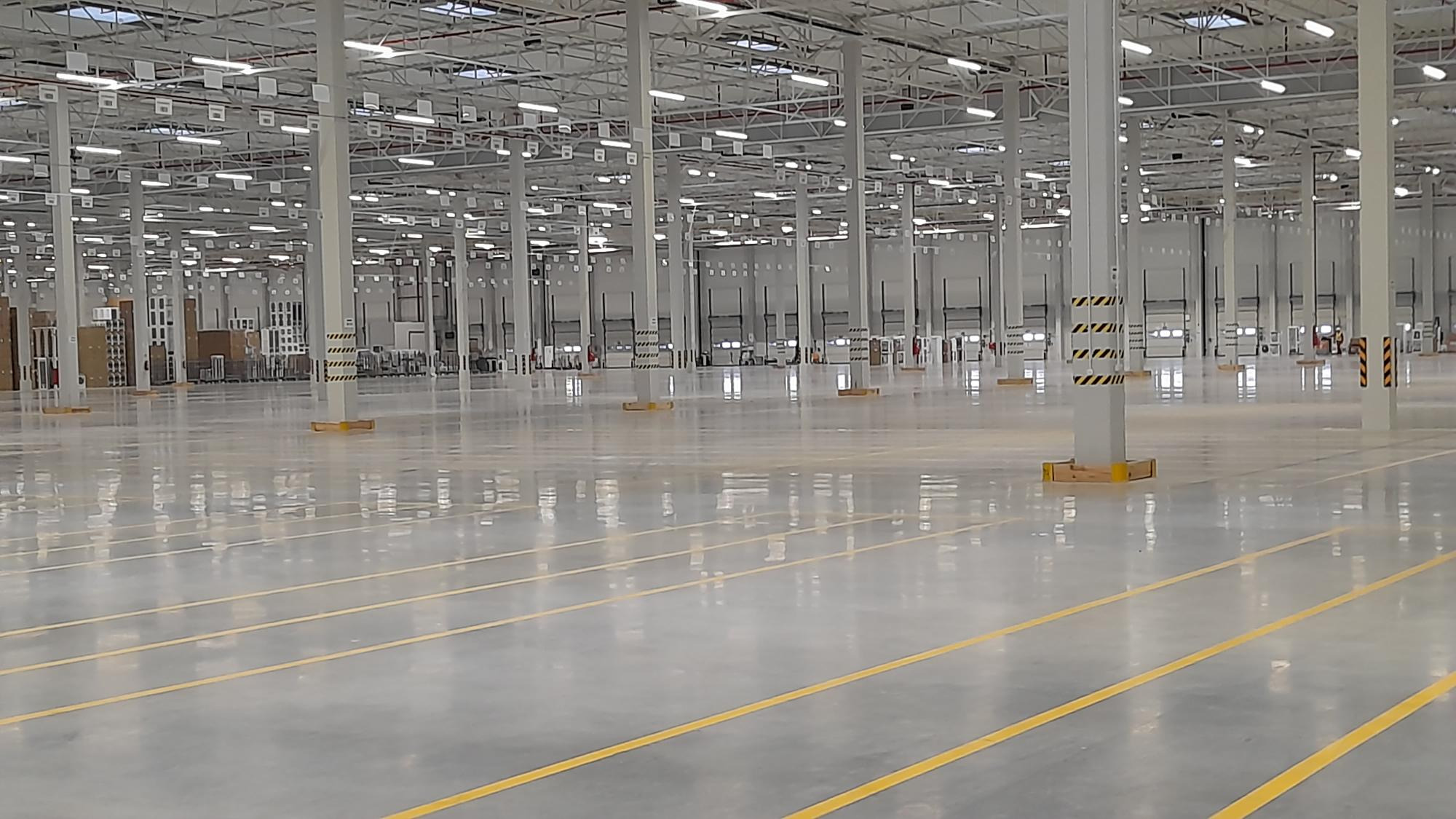Przestronne, puste wnętrze magazynu z polerowaną posadzką, żółtymi liniami wyznaczającymi strefy, wysokim sufitem z oświetleniem i rzędami kolumn z czarno-żółtymi pasami ostrzegawczymi u podstawy.