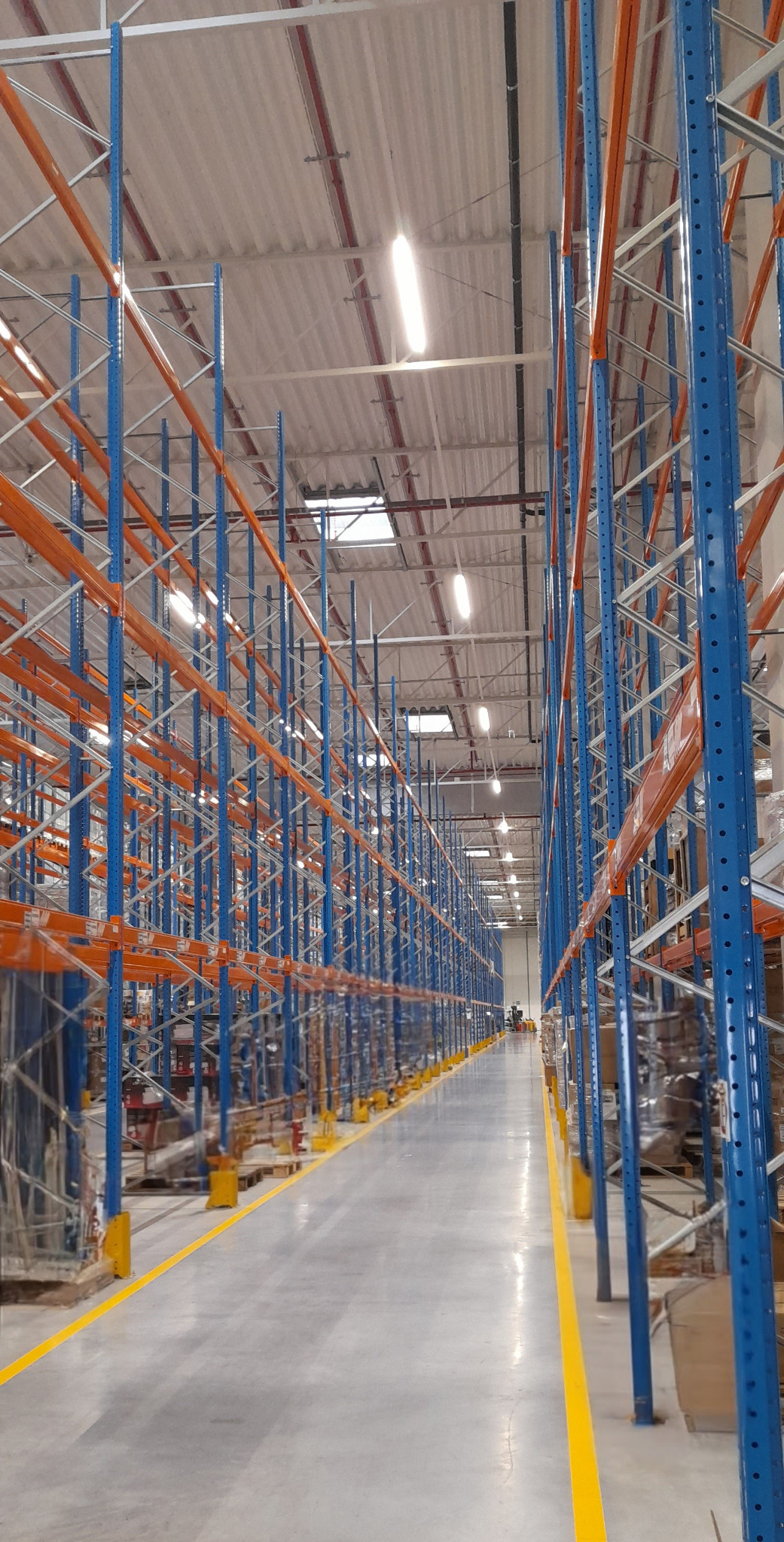 Długi korytarz w magazynie z regałami w kolorach niebieskim i pomarańczowym, widoczne palety z towarem i żółte linie na podłodze.