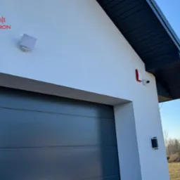 Zewnętrzny montaż kamery monitoringu z czujnikiem ruchu i sygnalizatorem, widoczny fragment elewacji domu z bramą garażową i czarnym dachem.