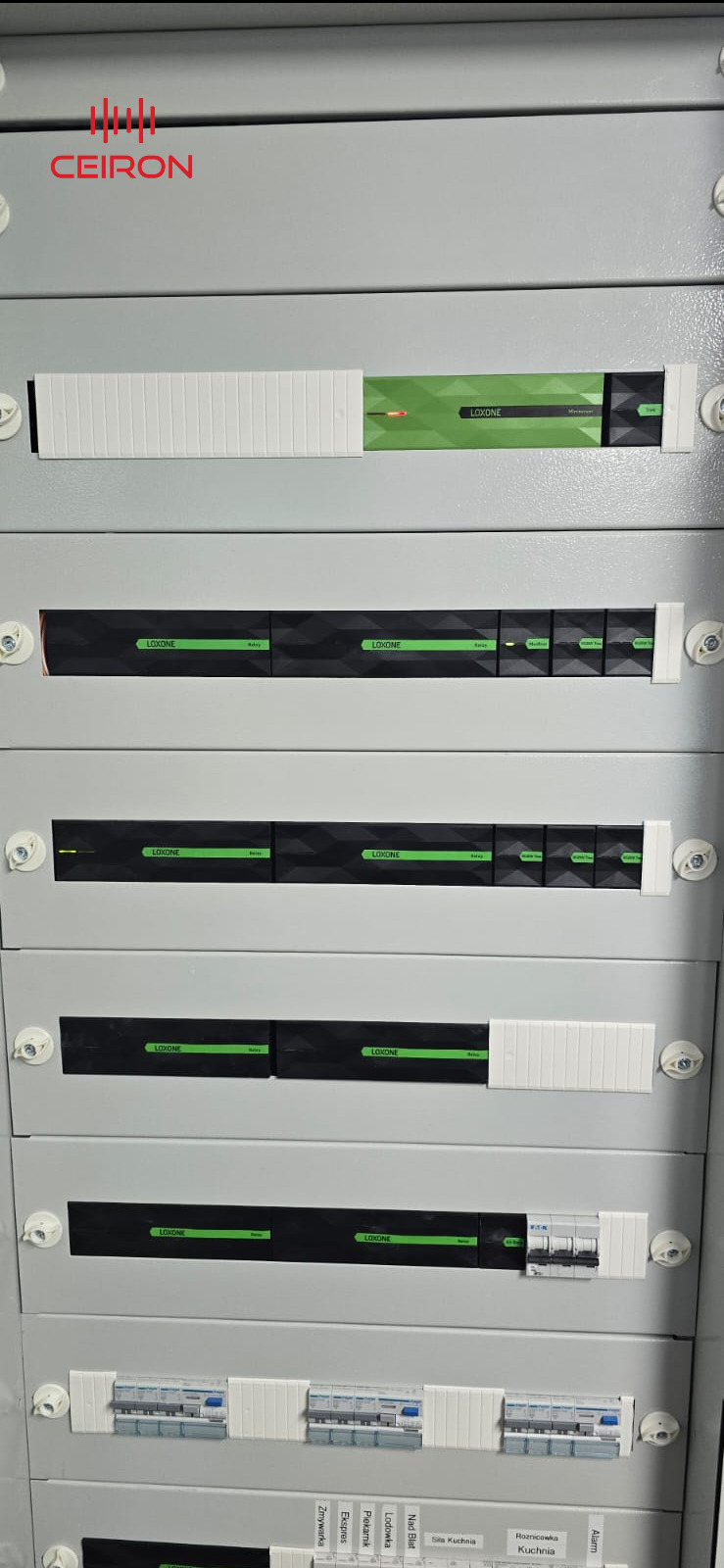 Rozdzielnia elektryczna z modułami Loxone, widoczne bezpieczniki i moduły sterujące systemem inteligentnego domu, szara obudowa z białymi elementami mocującymi.