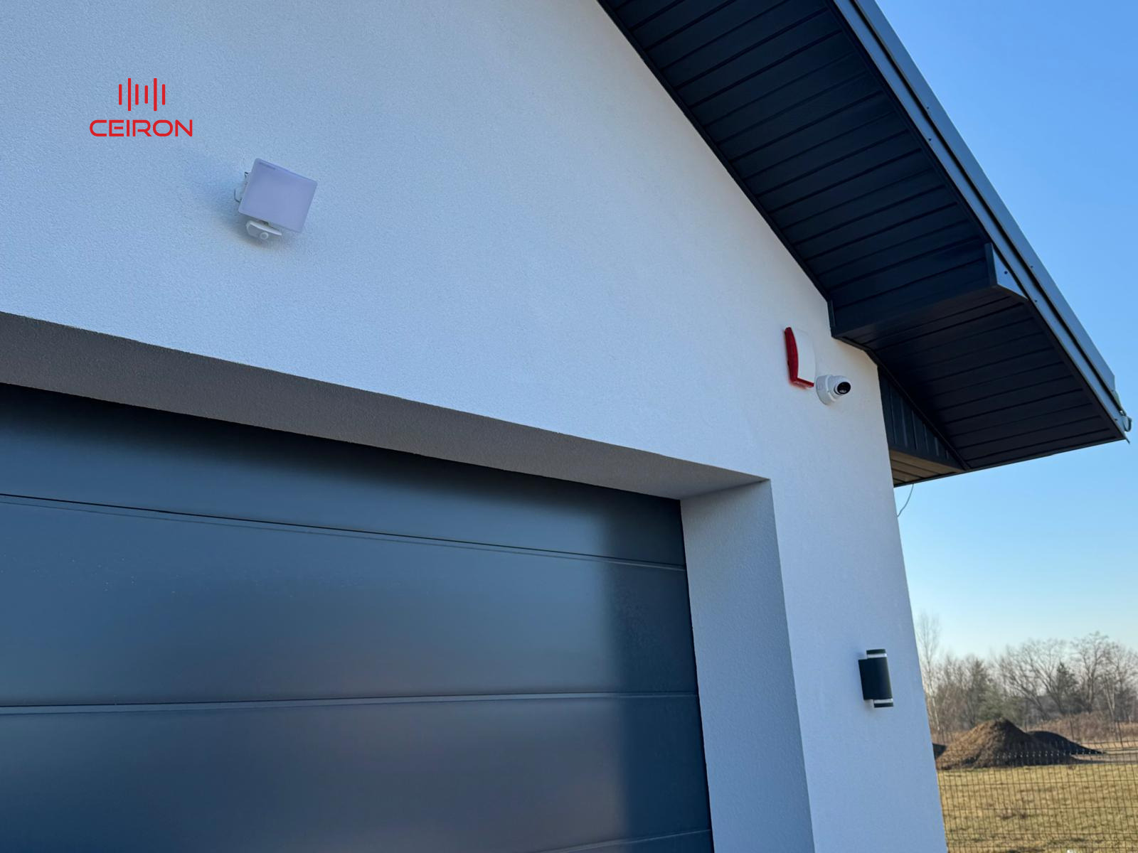Zewnętrzny montaż kamery monitoringu z czujnikiem ruchu i sygnalizatorem, widoczny fragment elewacji domu z bramą garażową i czarnym dachem.