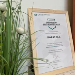 Oprawiony certyfikat 'Sprawdzony Partner w Biznesie' firmy Finari Sp. z o.o., z logo Crédit Agricole EFL Leasing, ustawiony obok dekoracyjnej rośliny, z widocznym protokołem przekazania...