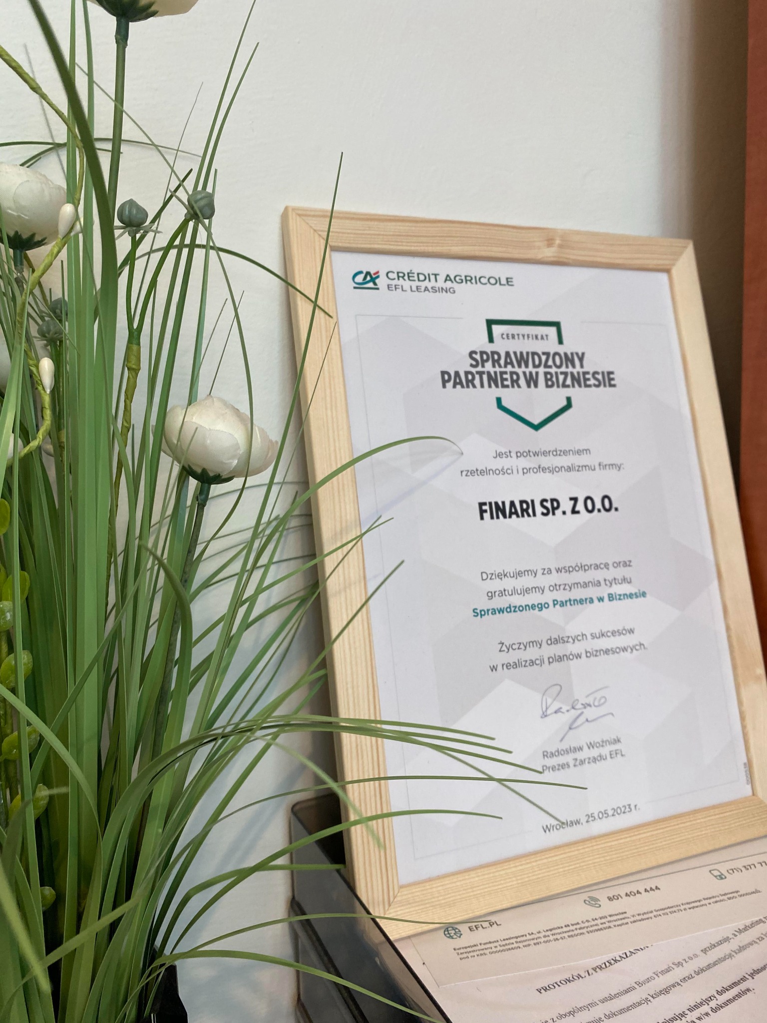 Oprawiony certyfikat 'Sprawdzony Partner w Biznesie' firmy Finari Sp. z o.o., z logo Crédit Agricole EFL Leasing, ustawiony obok dekoracyjnej rośliny, z widocznym protokołem przekazania...