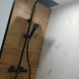 Pro-Mar kompleksowe wykończenie wnętrz - Czarny panel prysznicowy z deszczownicą i słuchawką, zamontowany na ścianie wyłożonej płytkami imitującymi drewno, sąsiadującej z płytkami marmurkowymi w jasnych odcieniach.
