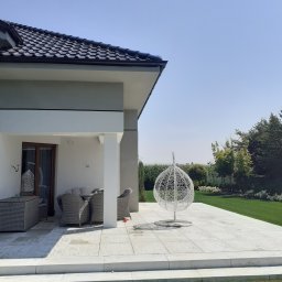 LIS-BUD BARTŁOMIEJ LISIECKI - Nowoczesny taras z jasnoszarą kostką, wiszącym fotelem kokonem i meblami ogrodowymi w tonacji szarości, widok na zadbany trawnik i zieleń.