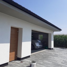 LIS-BUD BARTŁOMIEJ LISIECKI - Nowoczesny garaż z białym tynkiem strukturalnym, brązowymi drzwiami bocznymi i szarą kostką brukową, z widocznym samochodem osobowym wewnątrz i polem kukurydzy w tle.