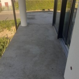 LIS-BUD BARTŁOMIEJ LISIECKI - Surowy betonowy taras z widocznymi śladami po pracach budowlanych, duże okno przesuwne z czarną ramą i biały filar, widok na ulicę i zieleń w tle.