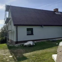 Dom jednorodzinny z nową, brązową dachówką i świeżo pomalowaną elewacją w odcieniach szarości, widoczne rusztowania przy ścianie szczytowej, trawnik z resztkami materiałów budowlanych.