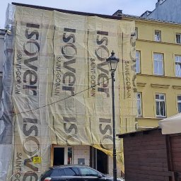 LIS-BUD BARTŁOMIEJ LISIECKI - Budynek w trakcie ocieplania, osłonięty siatką z nadrukiem Isover Saint-Gobain, widoczny fragment ulicy z zaparkowanym samochodem i fragmenty innych budynków.