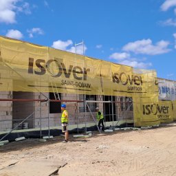 LIS-BUD BARTŁOMIEJ LISIECKI - Budowa domu jednorodzinnego z białego bloczka, zabezpieczona rusztowaniem i żółtą siatką z logo Isover, pracownicy w kamizelkach odblaskowych układają izolację termiczną.