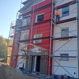 LIS-BUD BARTŁOMIEJ LISIECKI - Budynek wielorodzinny w trakcie prac elewacyjnych: część fasady z czerwonym tynkiem i rusztowaniami, pracownik w pomarańczowej kamizelce na rusztowaniu, druga część fasady z szarym tynkiem, w tle...