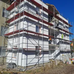 Ocieplanie ścian budynku z użyciem rusztowania, widoczni pracownicy w odblaskowych ubraniach, okna zabezpieczone folią, fragmenty starej elewacji widoczne obok nowej, ocieplonej ściany.