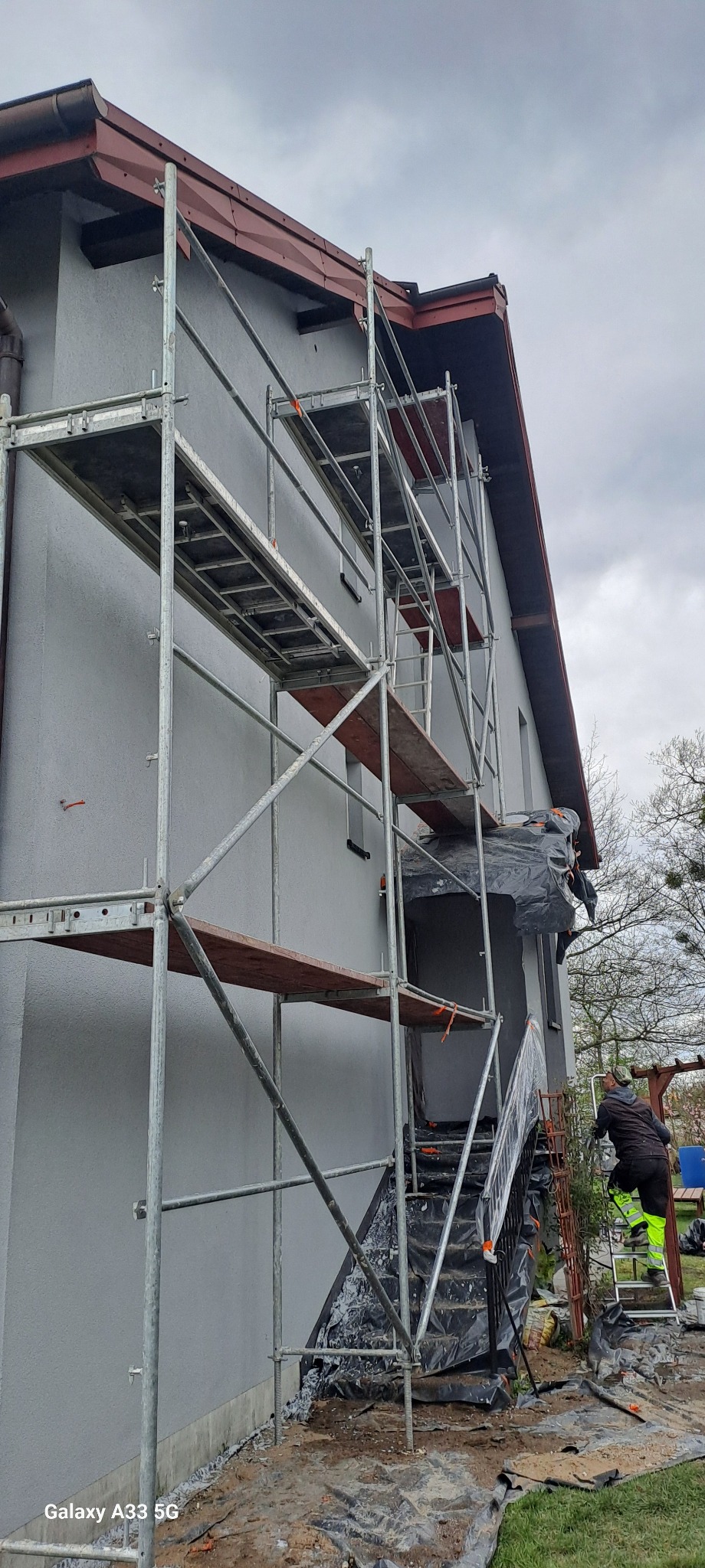 Dom jednorodzinny w trakcie prac elewacyjnych, widoczne rusztowania, zabezpieczone okna i drzwi folią, pracownik na drabinie w ubraniu roboczym.