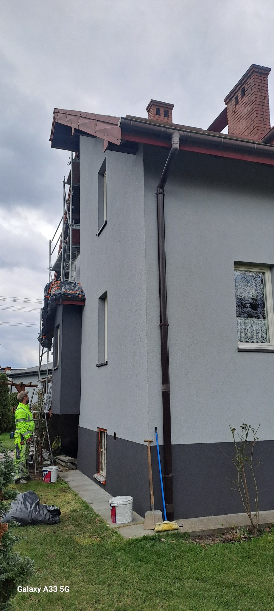 Dwukondygnacyjny dom z szarą elewacją, częściowo pomalowany na ciemniejszy odcień, widoczne rusztowania, pracownik w odblaskowym ubraniu, narzędzia malarskie i wiadro z farbą na trawniku, pochmurne...