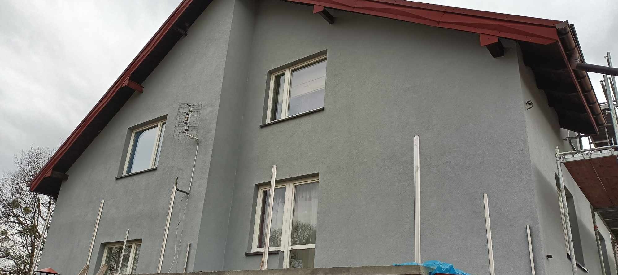 Szary dom jednorodzinny z nową elewacją, widoczna antena, rusztowanie i czerwona obróbka dachu.