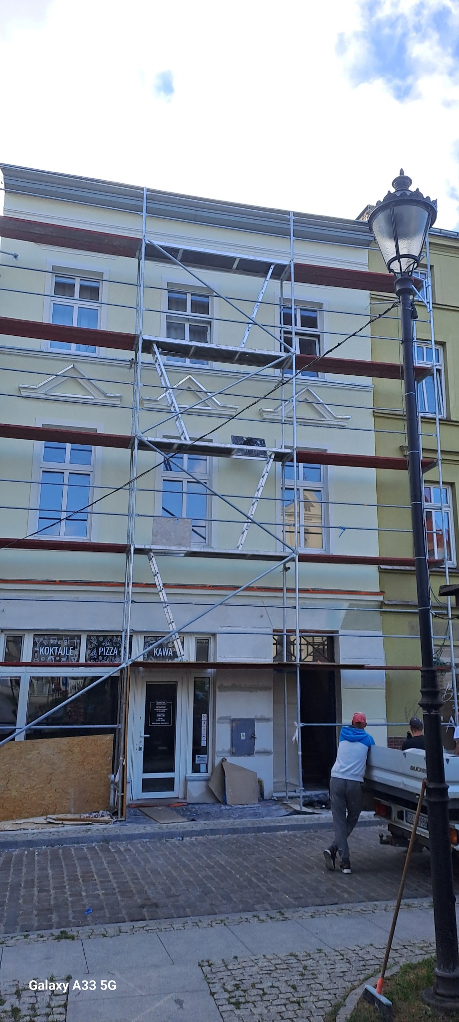 Budynek w trakcie renowacji elewacji, widoczne rusztowania, fragment odnowionej ściany i pracownicy przy samochodzie dostawczym.