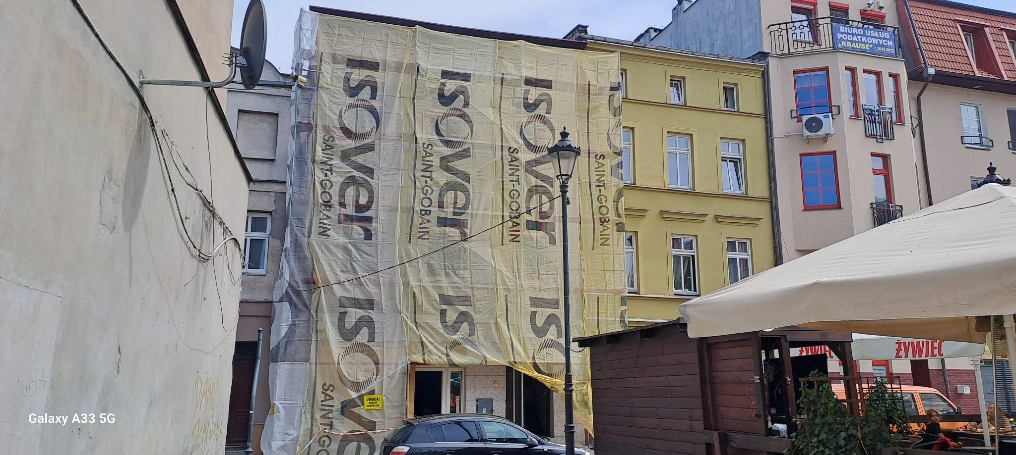 Budynek w trakcie ocieplania, osłonięty siatką z nadrukiem Isover Saint-Gobain, widoczny fragment ulicy z zaparkowanym samochodem i fragmenty innych budynków.