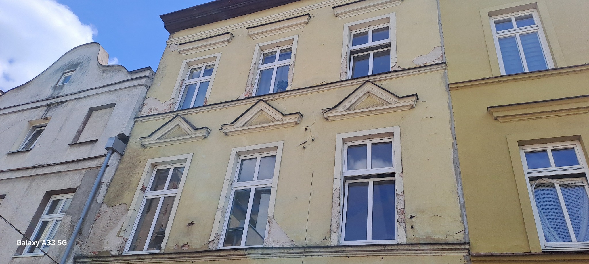 Żółta fasada kamienicy z widocznymi uszkodzeniami i pęknięciami, białe okna z ozdobnymi trójkątnymi elementami, sąsiedni budynek z szarą, zaniedbaną elewacją.