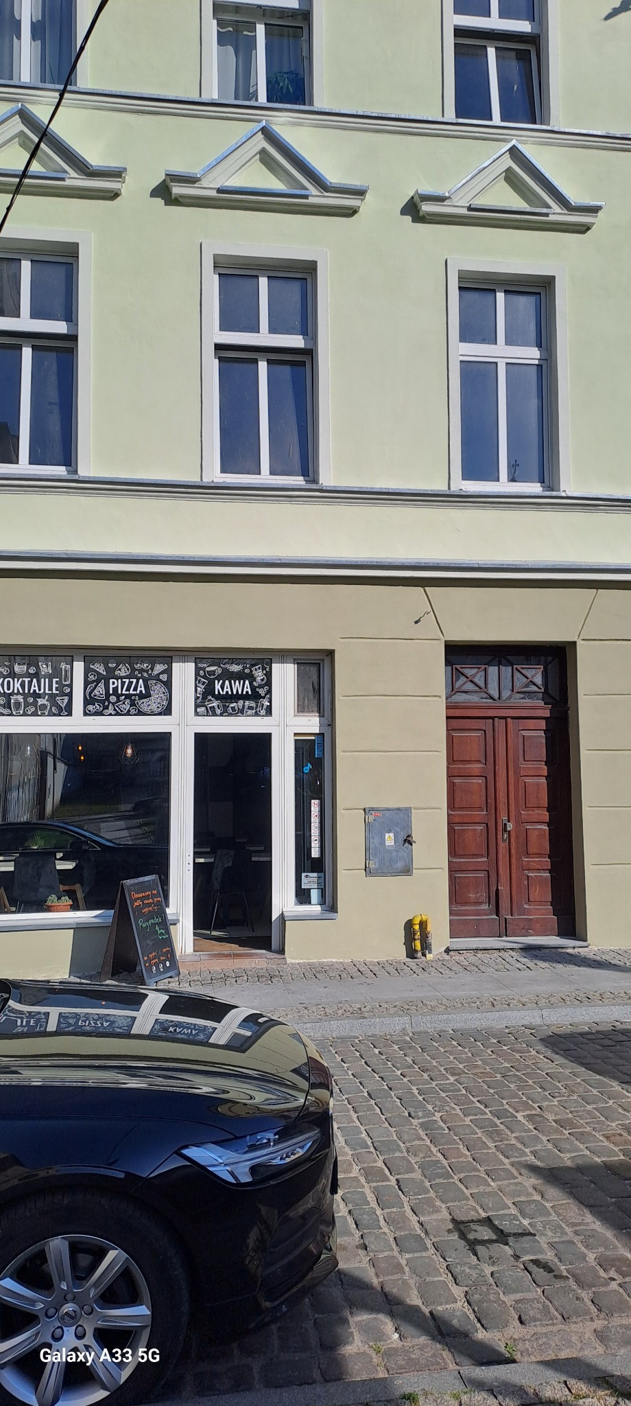 Zewnętrzny widok odnowionej elewacji budynku z restauracją, z oknami z białymi ramami i dekoracyjnymi elementami nad oknami, widoczny fragment czarnego samochodu na pierwszym planie, kostka brukowa...