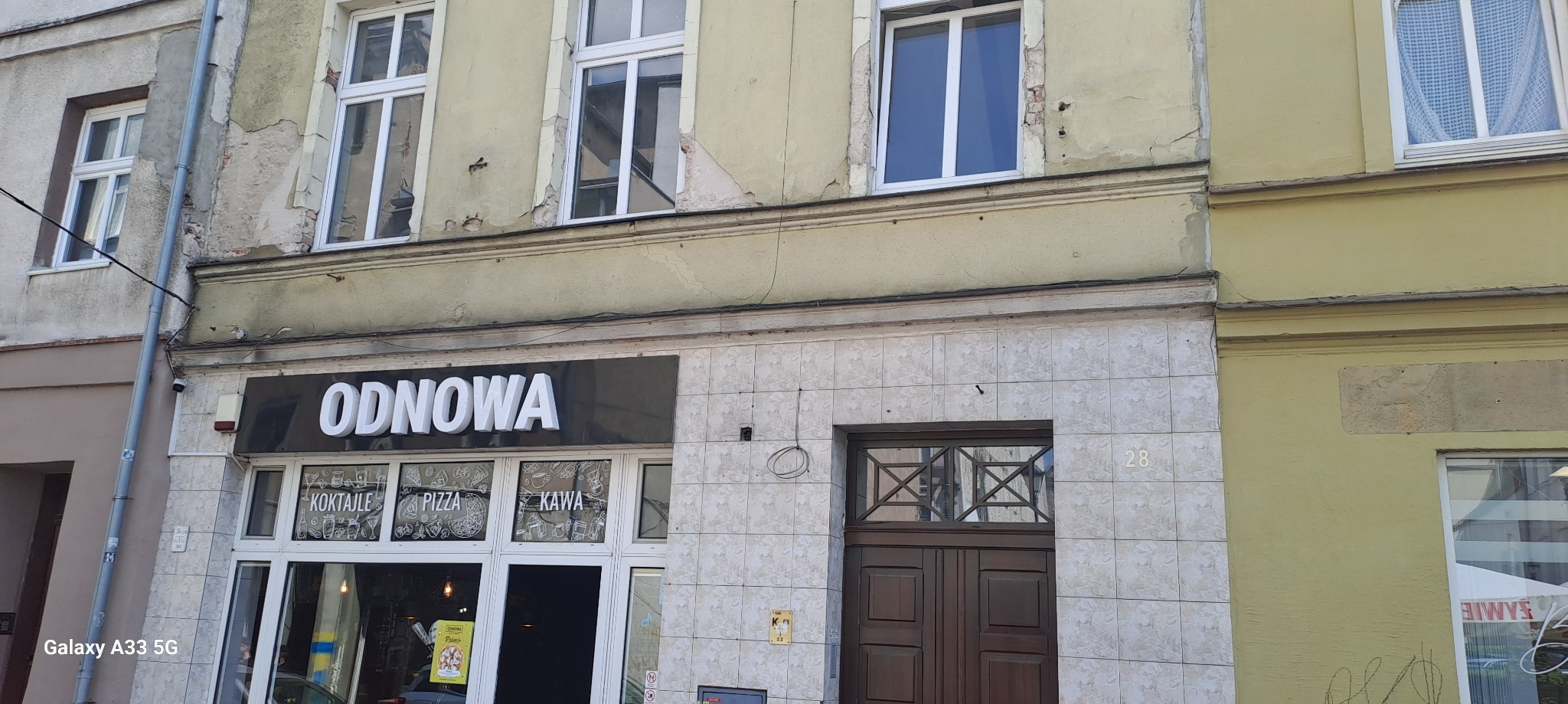 Fasada budynku z widocznymi śladami zużycia i odpadającą farbą, nad wejściem do lokalu z szyldem 'ODNOWA' - kontrast między zaniedbaną elewacją a nazwą lokalu.