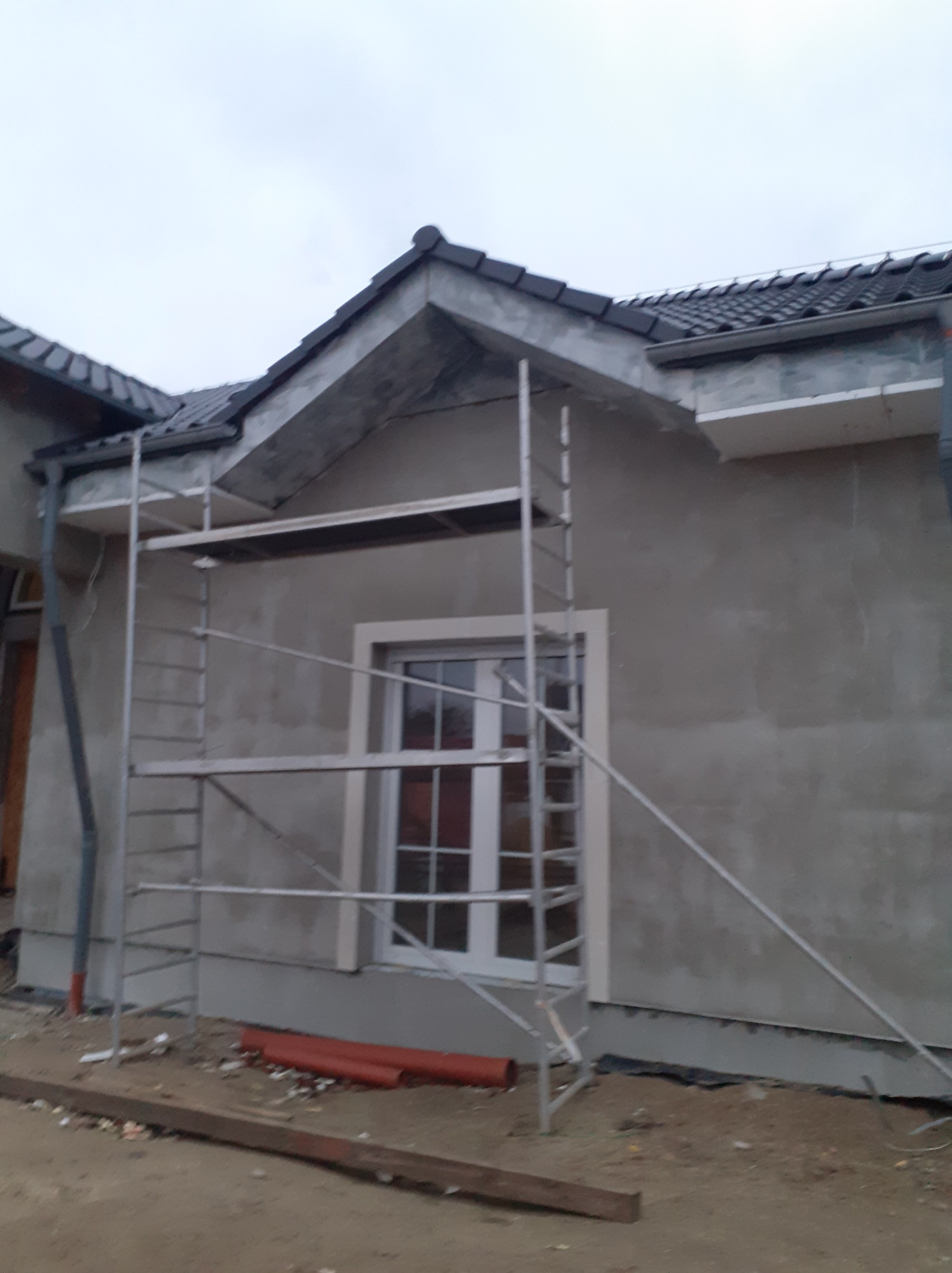 Dom jednorodzinny w trakcie ocieplania, widoczne rusztowanie, surowa elewacja z oknem, fragment dachu z czarną dachówką i rynną, teren budowy.