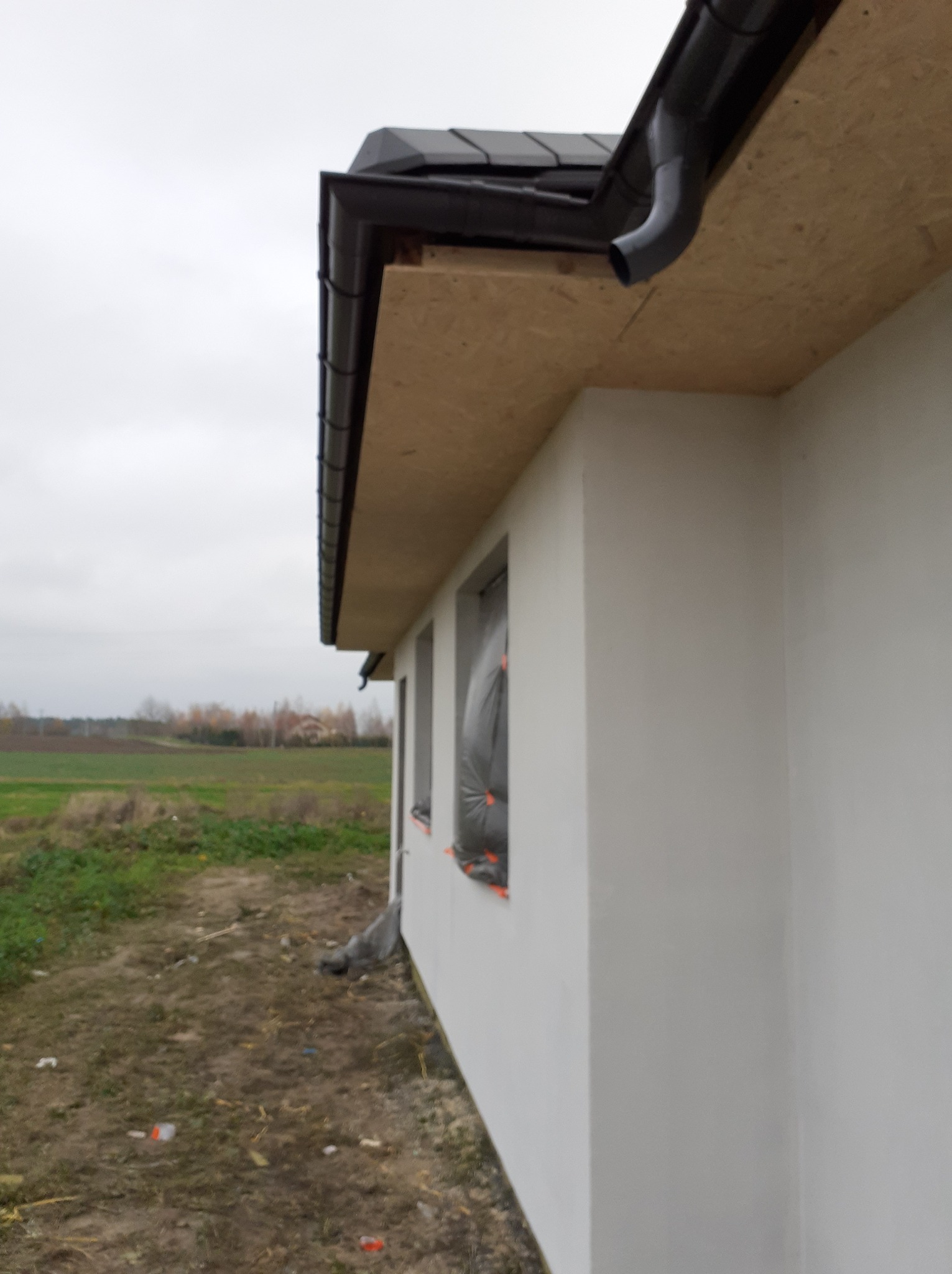 Fragment ocieplonego domu jednorodzinnego z zamaskowanymi oknami, widoczne orynnowanie i płyta OSB pod dachem, w tle pola i drzewa.