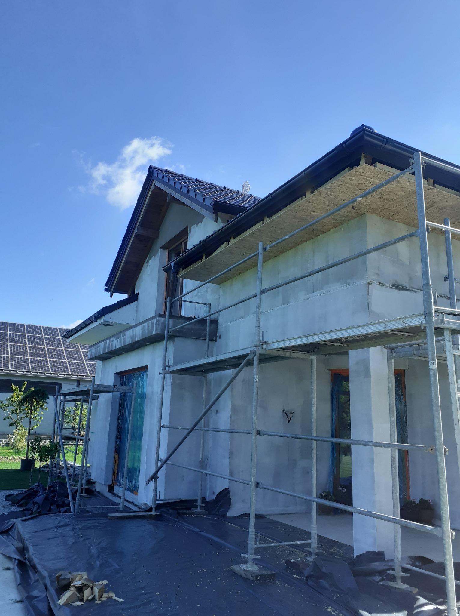 Dom jednorodzinny w trakcie ocieplania, widoczne rusztowania, zabezpieczone okna folią, częściowo otynkowana elewacja, w tle panele słoneczne na dachu sąsiedniego budynku.