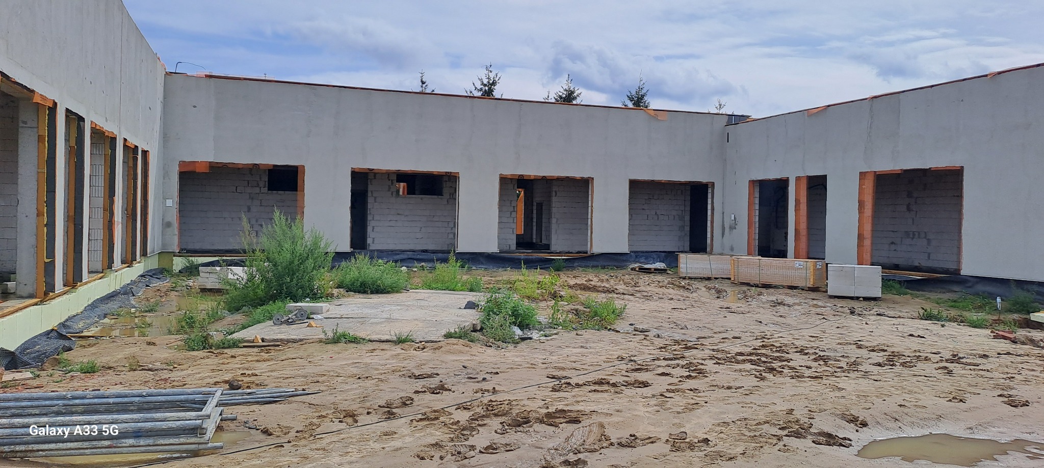 Budynek w trakcie ocieplania ścian zewnętrznych, widoczne pakiety materiałów izolacyjnych, niezadaszony teren budowy z piaskiem i kałużami.