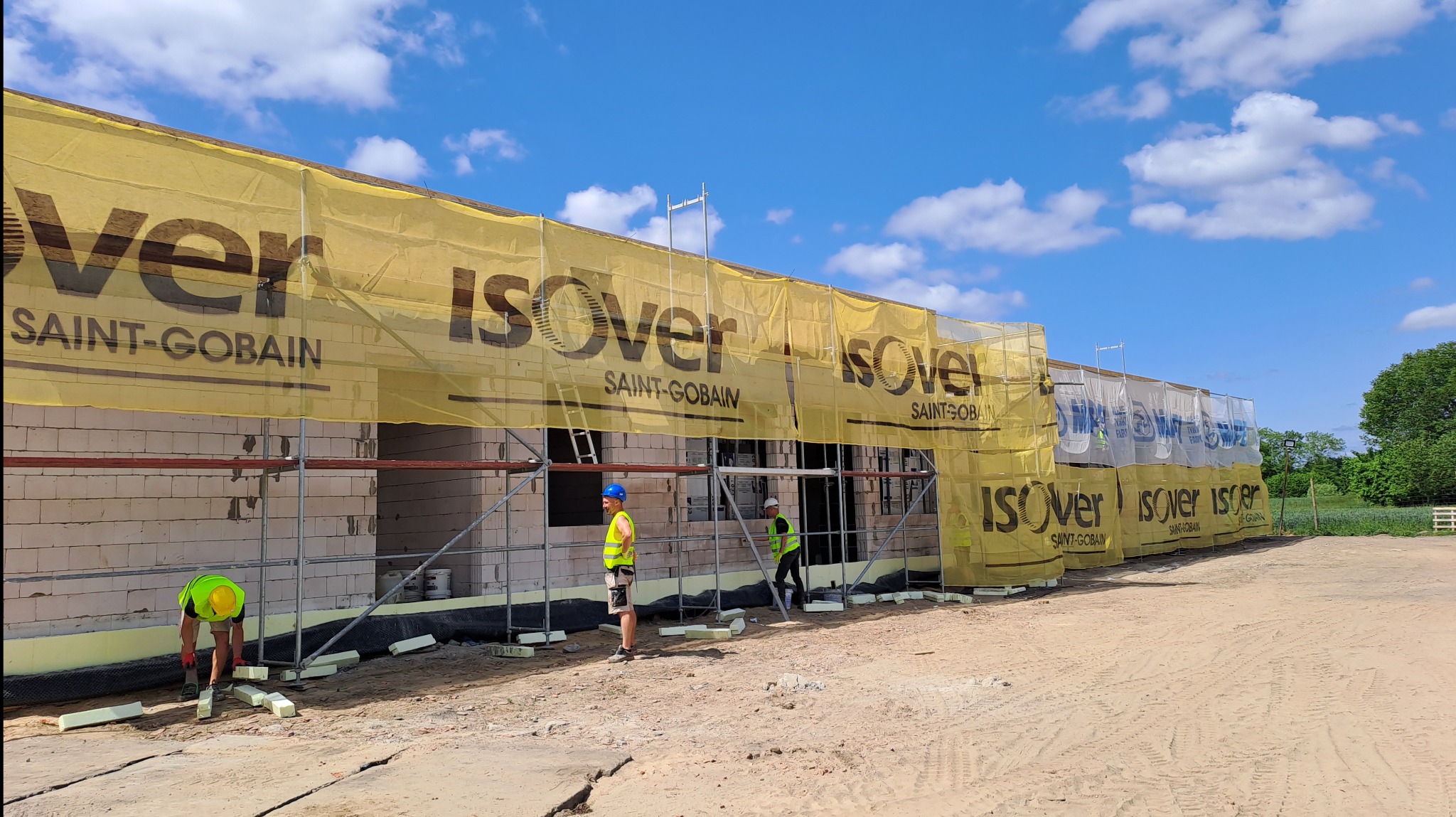 Budowa domu jednorodzinnego z białego bloczka, zabezpieczona rusztowaniem i żółtą siatką z logo Isover, pracownicy w kamizelkach odblaskowych układają izolację termiczną.
