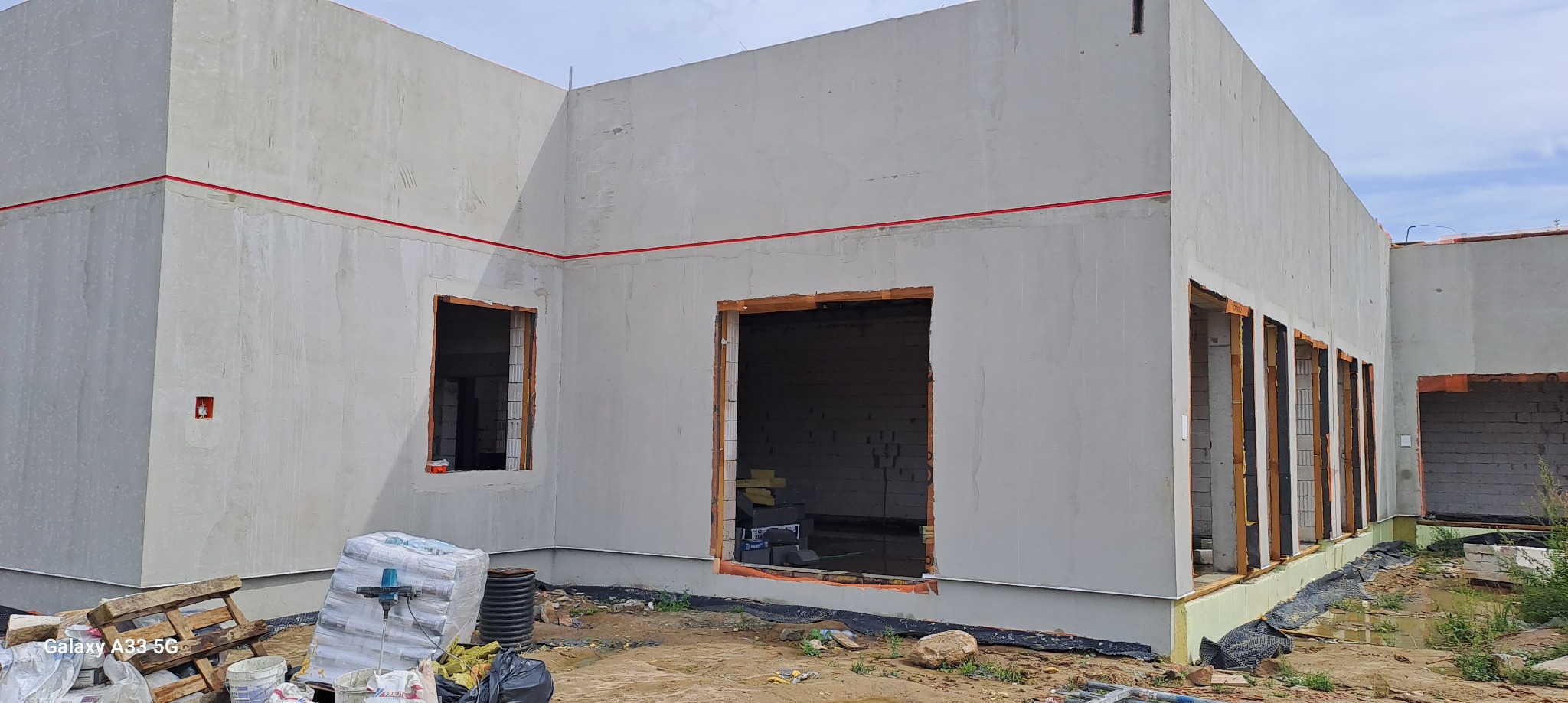 Surowy stan budynku jednorodzinnego z widocznymi otworami okiennymi i zaznaczoną linią poziomą na elewacji, materiały budowlane na palecie w pobliżu.