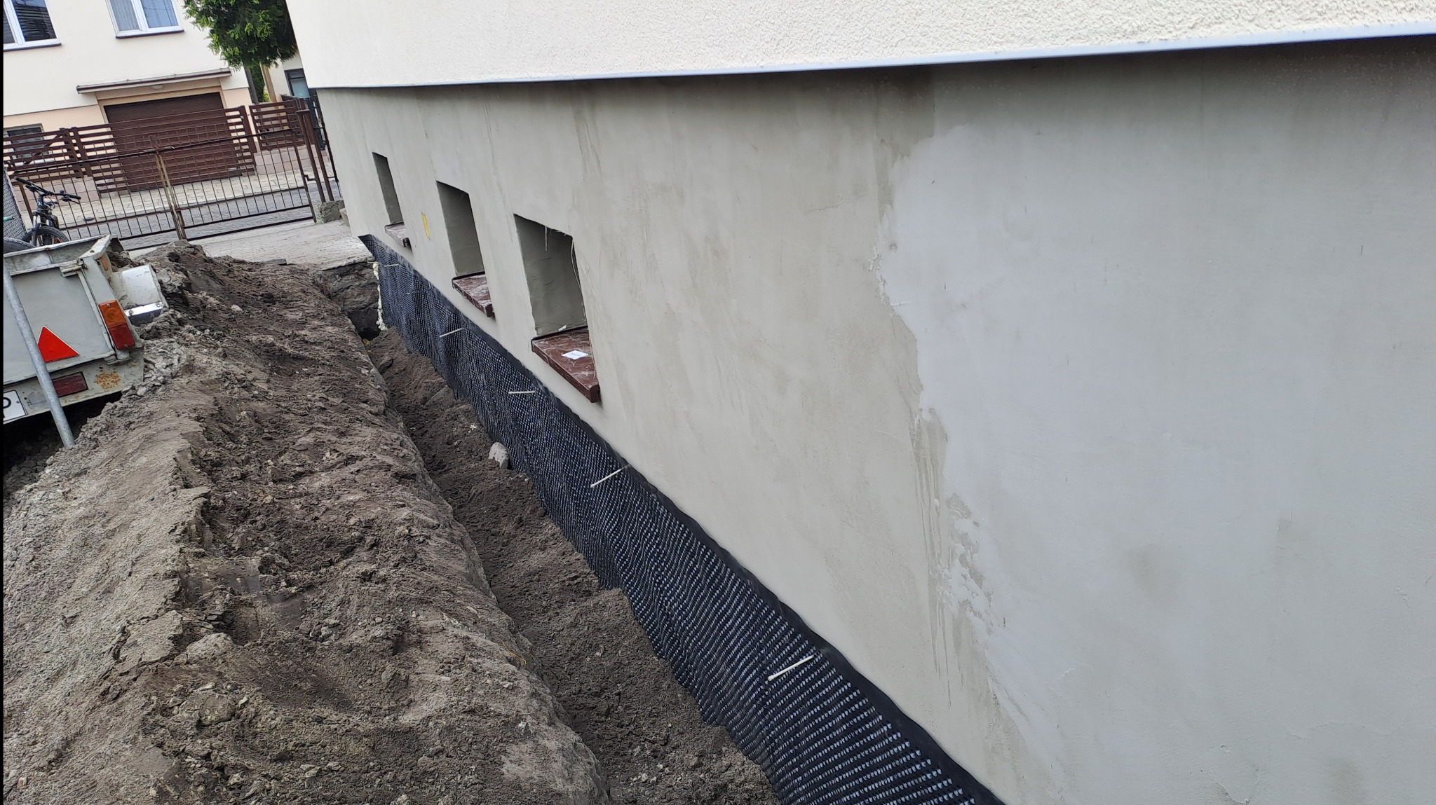 Przygotowanie fundamentów budynku: wykop wzdłuż ściany, zabezpieczona folia kubełkowa, widoczne okienka piwniczne. Ściana z surowym tynkiem. Przyczepka samochodowa w tle.