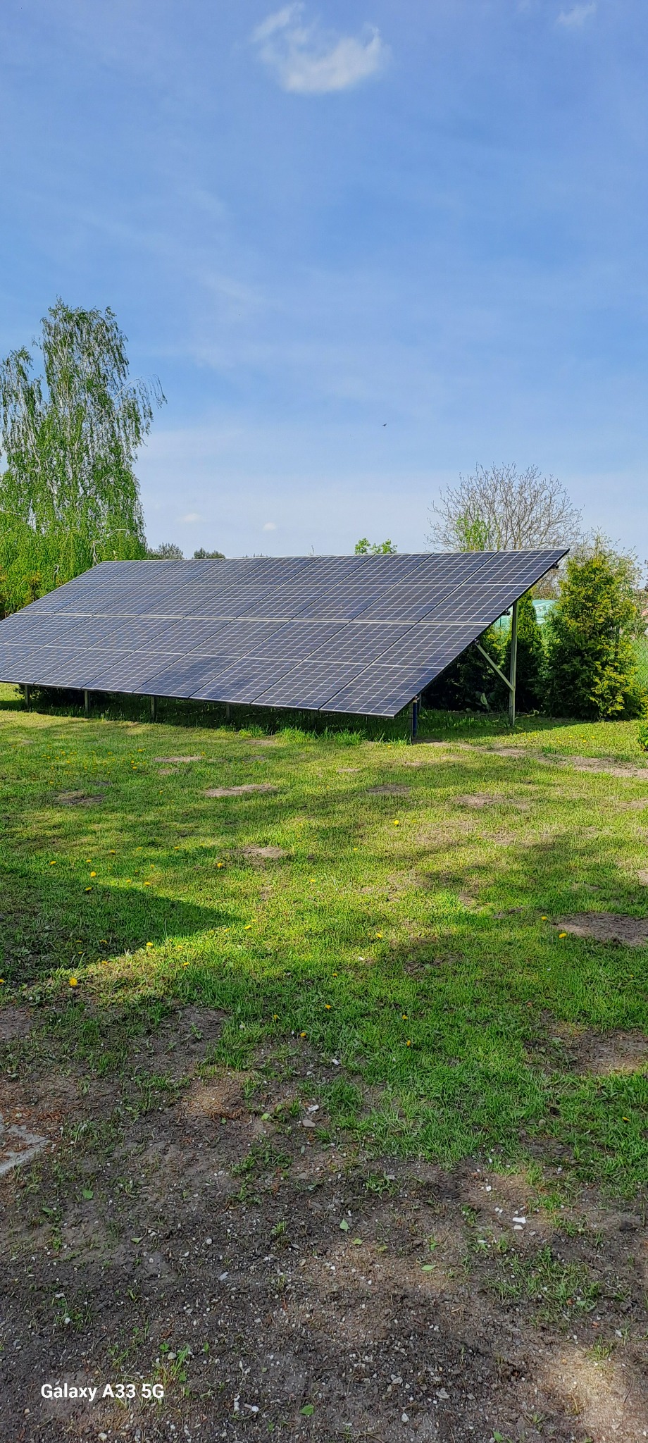 Panel słoneczny na metalowej konstrukcji, umieszczony na trawniku z widocznymi śladami ziemi, w tle drzewa i błękitne niebo.