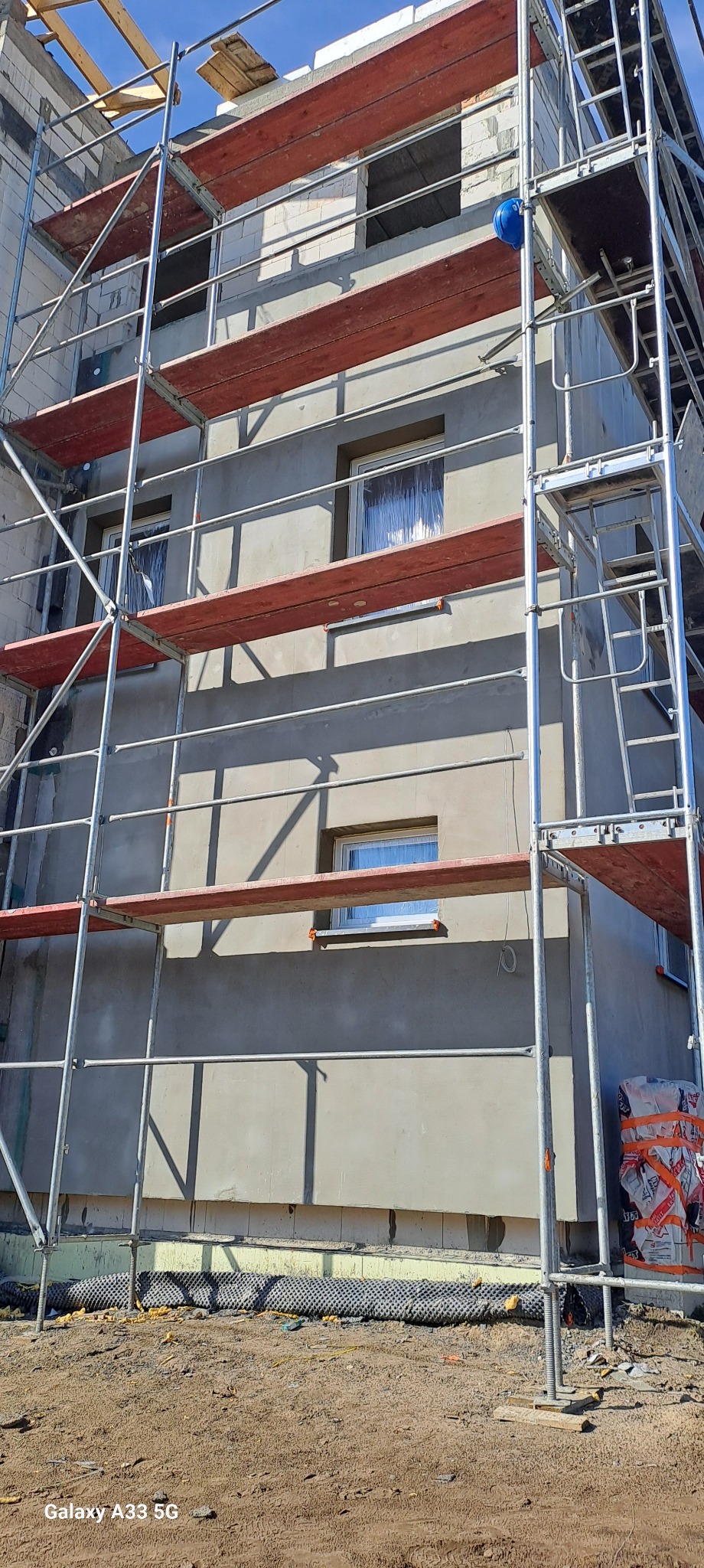 Budynek w Grudziądzu w trakcie ocieplania, widoczne rusztowania i częściowo otynkowana elewacja, okna zabezpieczone folią.