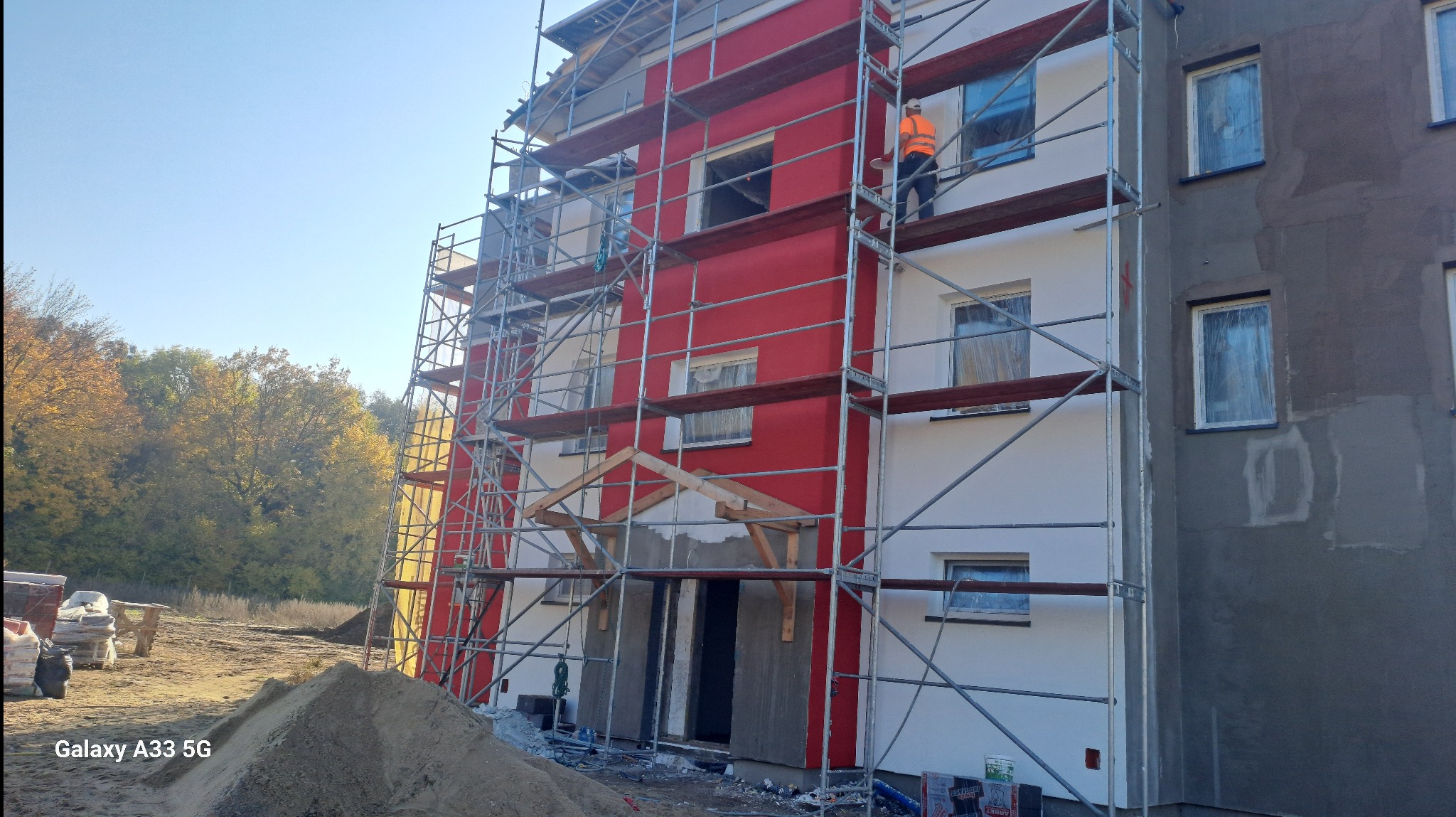 Budynek wielorodzinny w trakcie prac elewacyjnych: część fasady z czerwonym tynkiem i rusztowaniami, pracownik w pomarańczowej kamizelce na rusztowaniu, druga część fasady z szarym tynkiem, w tle...