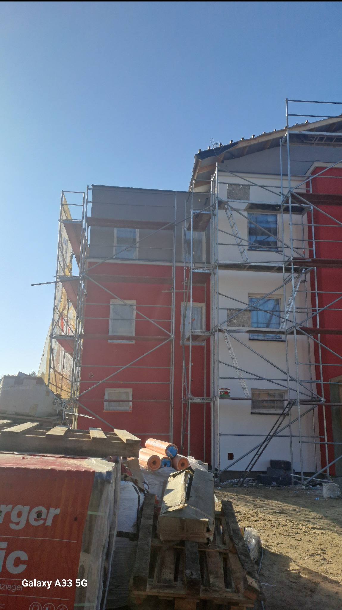 Wielopiętrowy budynek w trakcie ocieplania, widoczne rusztowania, częściowo otynkowana biała ściana i czerwona ściana, na pierwszym planie materiały budowlane.