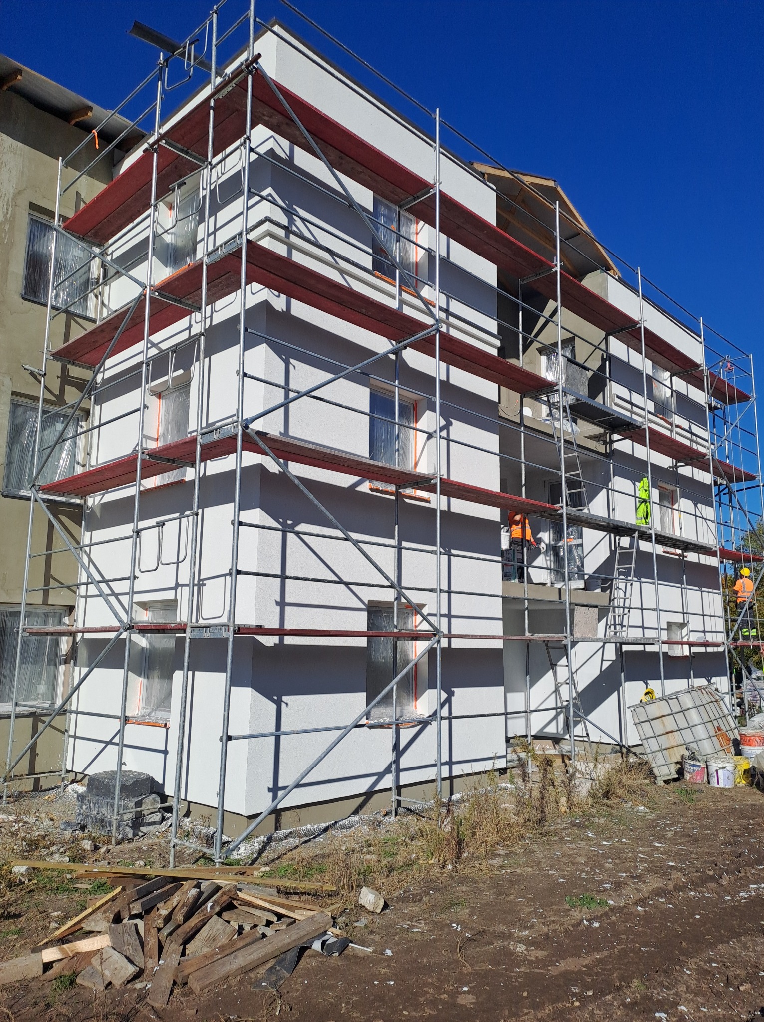 Ocieplanie ścian budynku z użyciem rusztowania, widoczni pracownicy w odblaskowych ubraniach, okna zabezpieczone folią, fragmenty starej elewacji widoczne obok nowej, ocieplonej ściany.