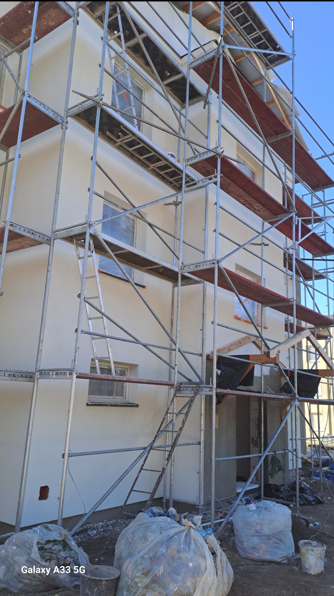 Biała fasada domu w trakcie prac wykończeniowych, z widocznymi rusztowaniami, zabezpieczonymi oknami folią i workami z odpadami budowlanymi u podstawy.