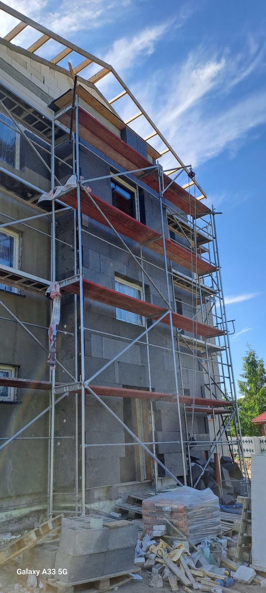 Budynek w trakcie ocieplania, widoczne rusztowanie, szare płyty izolacyjne, fragmenty zaprawy i materiałów budowlanych na ziemi, błękitne niebo w tle.
