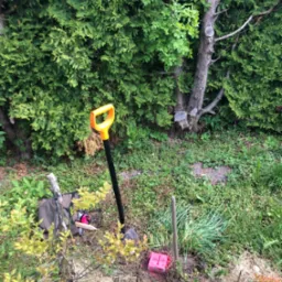 Sonda geodezyjna pomalowana na różowo wbita w ziemię obok łopaty z żółtym uchwytem, na tle zieleni. Widoczne narzędzia pomiarowe i torba. Prace terenowe.