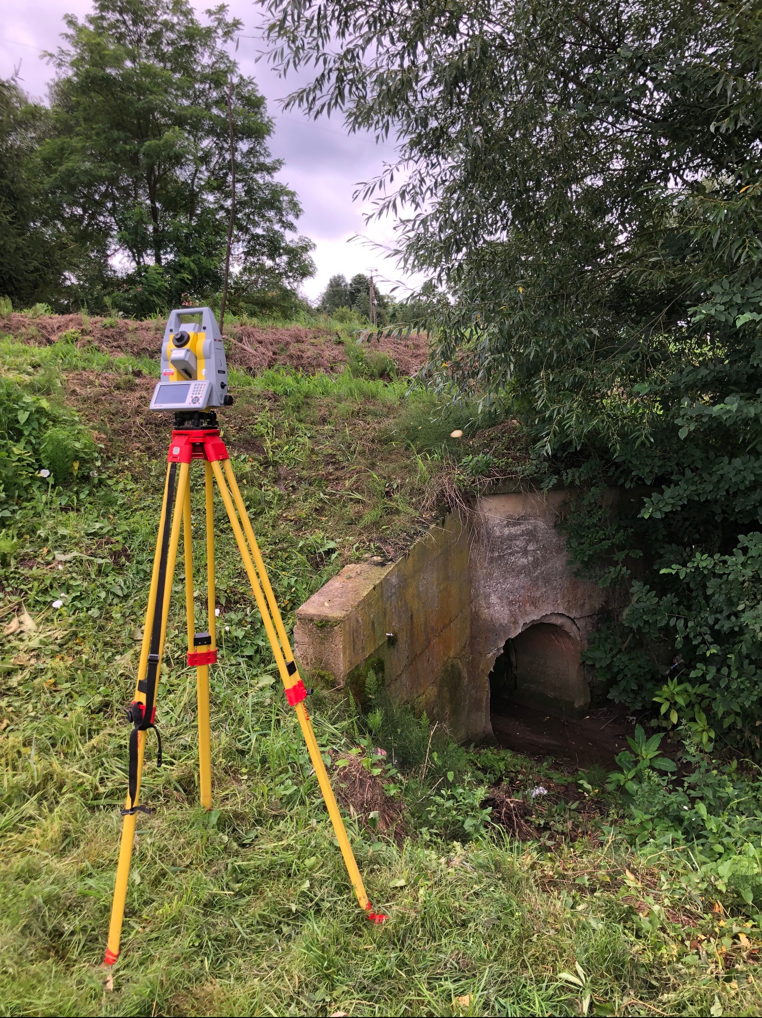 Żółty teodolit na trójnogu ustawiony przed betonowym przepustem w ziemnym nasypie, zarośniętym trawą i krzewami. Pomiar geodezyjny w terenie.