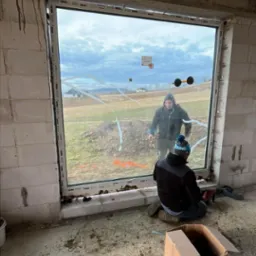 Dwóch mężczyzn instaluje duże okno w surowym stanie, widoczne przyssawki do szkła, narzędzia i karton na podłodze.