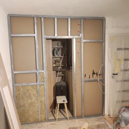 SpecINSTAL Adrian Strugała - Wnętrze w trakcie tworzenia ścianki działowej z metalowym szkieletem, wypełnionym wełną mineralną, z widocznym otworem drzwiowym i instalacją elektryczną, na podłodze resztki materiałów budowlanych...