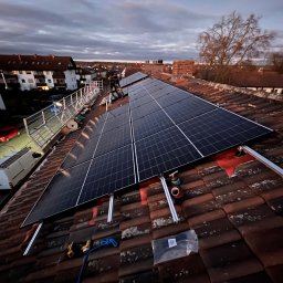 SpecINSTAL Adrian Strugała - Panele słoneczne w trakcie montażu na dachu z czerwonej dachówki, widoczne narzędzia i zabezpieczenia, rusztowanie w tle, pochmurne niebo.