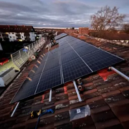 Panele słoneczne w trakcie montażu na dachu z czerwonej dachówki, widoczne narzędzia i zabezpieczenia, rusztowanie w tle, pochmurne niebo.